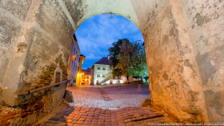 View through a rough masonry arch with exposed stone and brick toward a warmly lit cobbled square and colorful buildings at dusk.