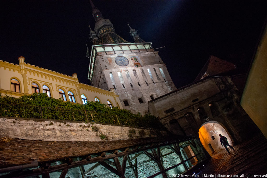 Tilted night view of Sighisoara Clock Tower above stone walls, timber gallery, and a glowing arched passage.