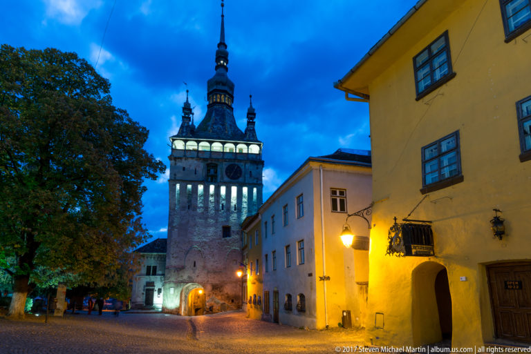 Sighisoara Clock Tower illuminated above a cobbled street at blue hour, with warm lamps, gateway arch, and neighboring historic buildings.