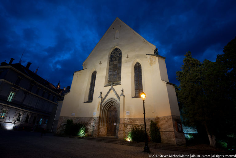 Gothic church façade at blue hour with pointed doorway, tall lancet windows, large central window, pale plaster, and masonry base.
