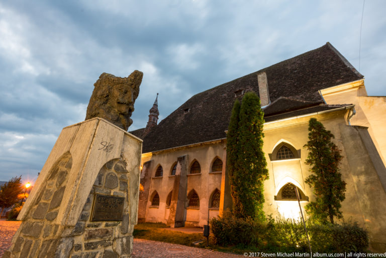 Stone memorial beside an illuminated historic church with pointed Gothic windows, ivy-covered walls, and a steep dark tiled roof.