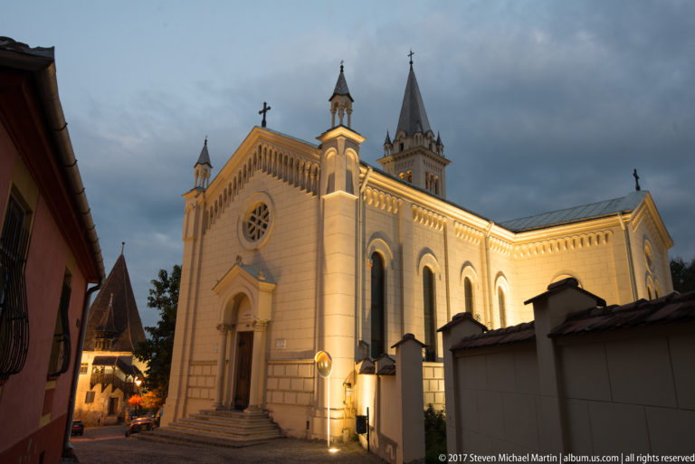 Oblique dusk view of an illuminated pale church with pointed entrance, circular window, arcading, and tall bell tower behind.