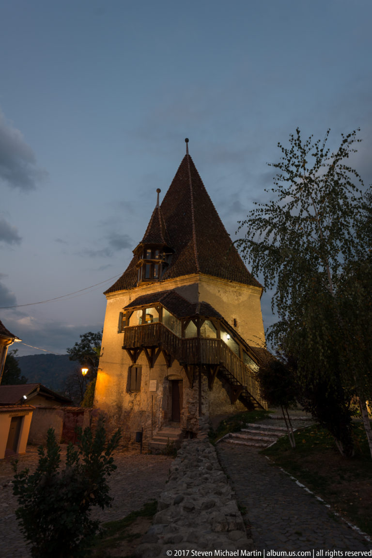 Illuminated historic masonry structure at dusk with a broad steep tiled roof and projecting timber gallery above a rough stone base.