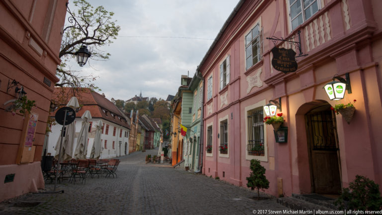 Narrow cobblestone street lined with pink, green, orange, and white historic houses, hanging signs, lanterns, and café tables.