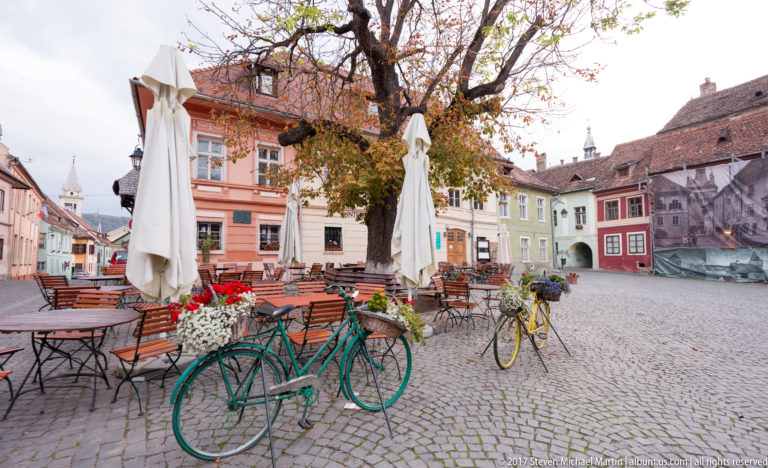 Cobbled Sighisoara square with café tables, closed umbrellas, flower planters, decorative bicycles, and pastel historic buildings.