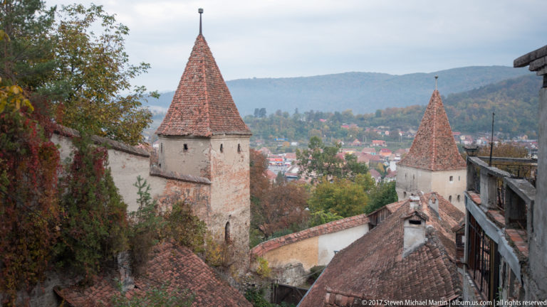 Two stone defensive towers with steep red-tiled roofs along Sighisoara’s fortified wall, town and wooded hills beyond.