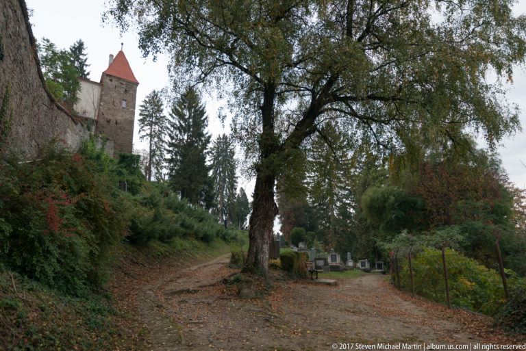 Leaf-covered cemetery path with grave markers and mature trees below a stone defensive tower with a red roof.