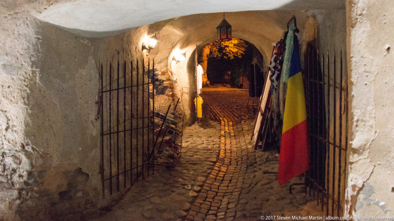 Narrow cobbled arched passage with iron grilles, wall lamps, and a Romanian flag hanging at right.