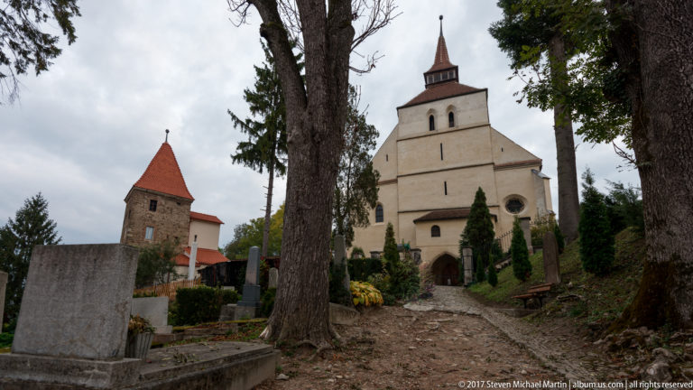 Stone path leading to the Church on the Hill, with its pale stepped façade and a rough masonry tower with red pyramidal roof at left.