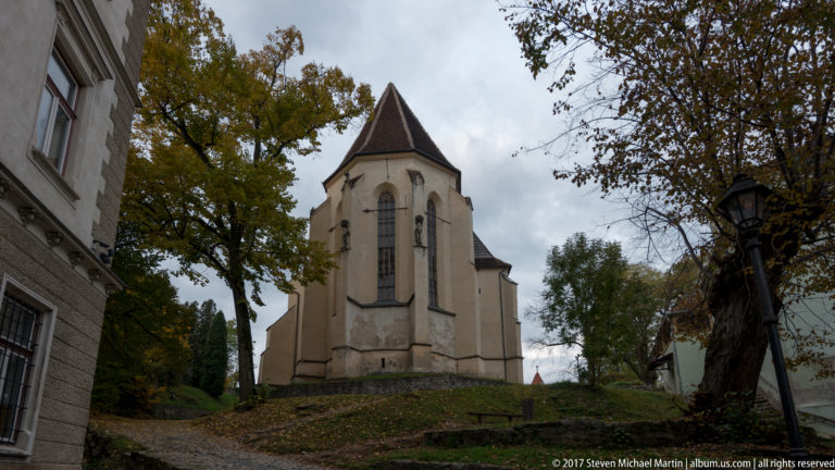 Church on the Hill seen from below, with pale walls, tall Gothic windows, steep roof, trees, and sloping ground.