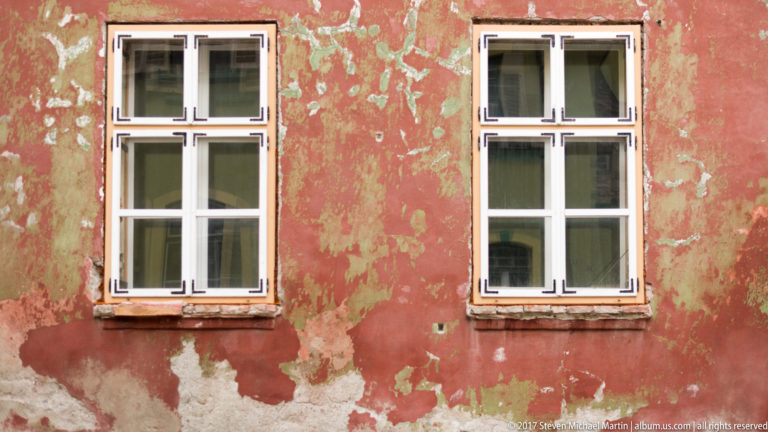 Two white-framed windows in a red plaster wall with extensive peeling paint and exposed weathered surface.