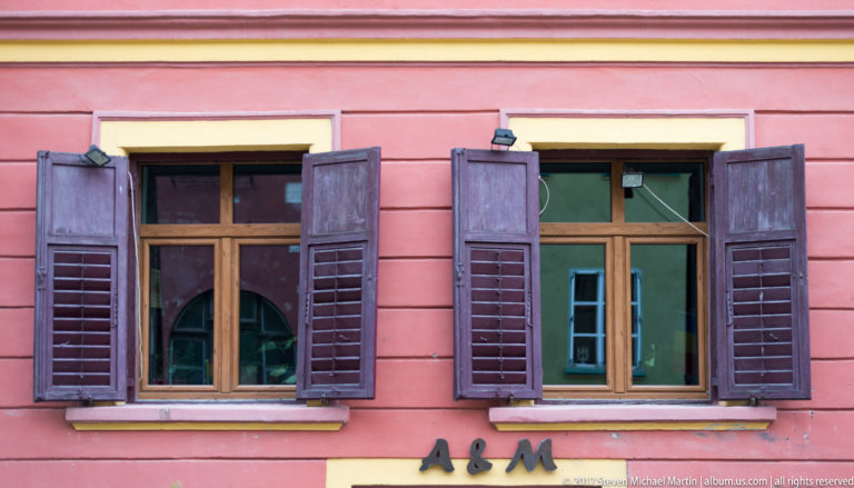 Two tall wood-framed windows with open dark shutters set in a pink plaster façade with cream trim.