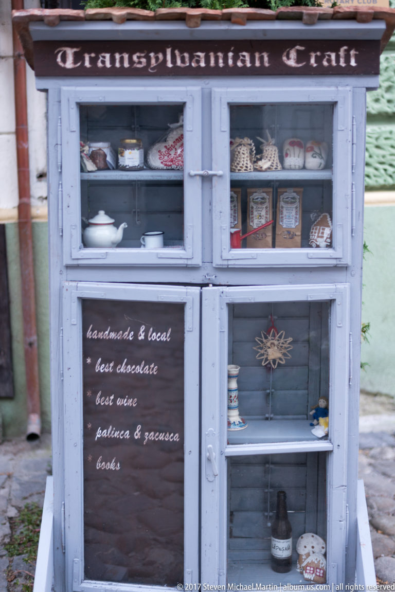 Painted street cabinet filled with ceramics, crafts, bottles, and souvenirs beside a chalkboard listing handmade local products.