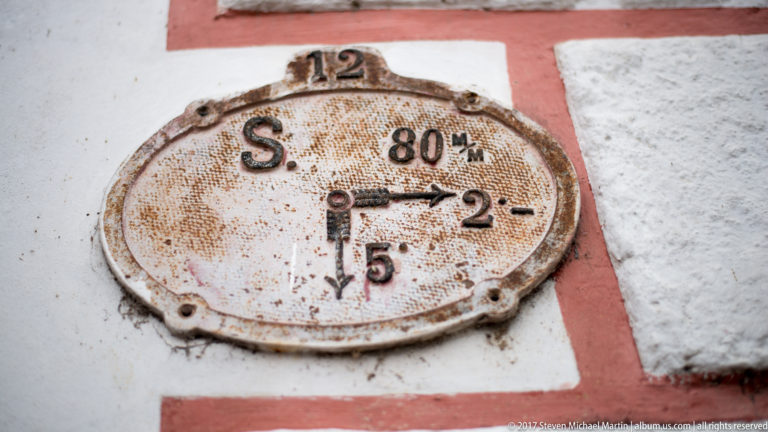 Rust-speckled oval metal utility plate with raised letters, numbers, and diagram markings mounted on a red-and-white plaster wall.