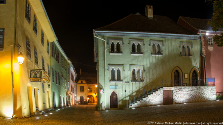Historic pale building with pointed-arch windows and exterior stairs on an illuminated cobbled street at night.