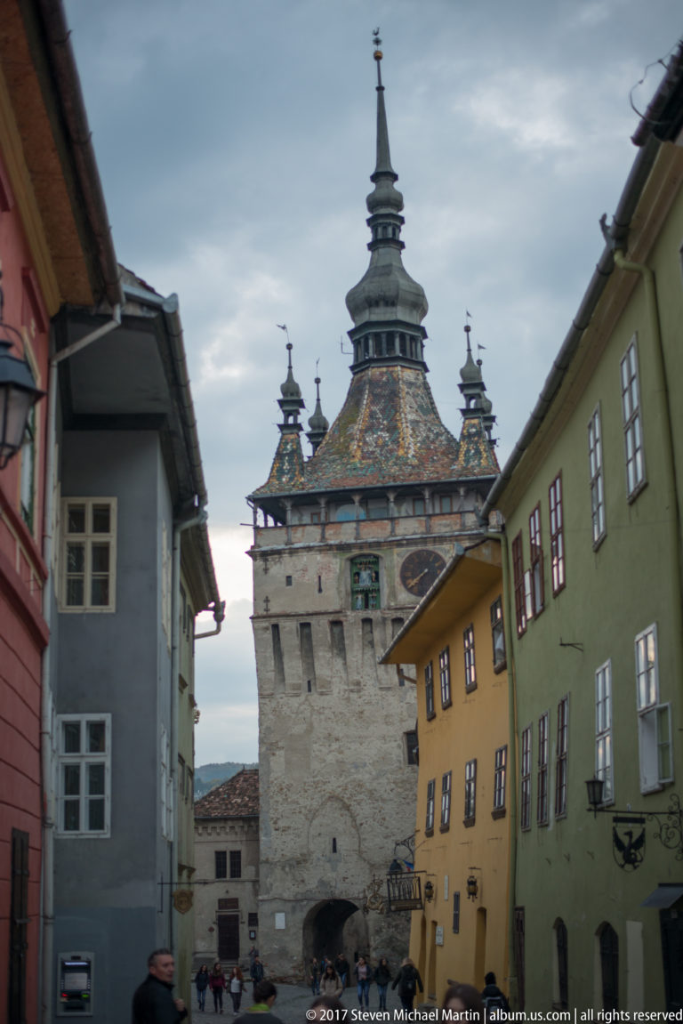 Sighisoara Clock Tower at the end of a narrow street lined by gray, yellow, and green historic buildings, with pedestrians near its base.
