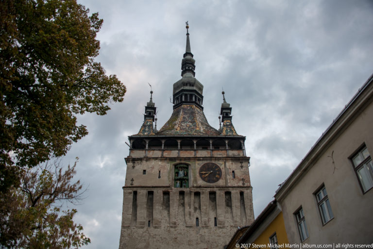 Low-angle view of Sighisoara Clock Tower with stone façade, clock face, polychrome roof, corner turrets, and central spire.