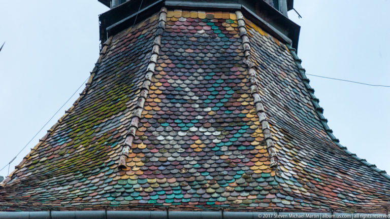Close-up of overlapping multicolored glazed tiles and moss on the steep roof of Sighisoara’s Clock Tower.