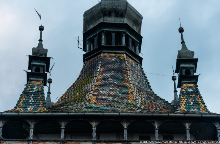 Close view of the Sighisoara Clock Tower roof with multicolored glazed tiles, central lantern, four corner turrets, and patches of moss.
