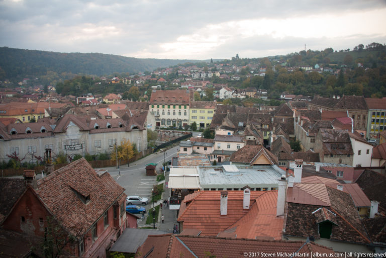 Elevated view across Sighisoara’s red-tiled roofs, streets, and larger buildings toward residential hills under an overcast sky.