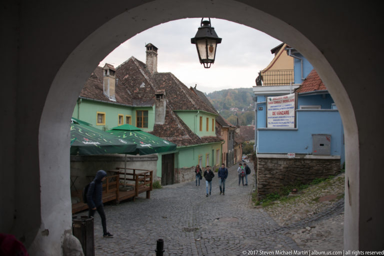 Cobblestone street and pedestrians seen through a large masonry arch with a hanging lantern, pastel houses lining the lane beyond.