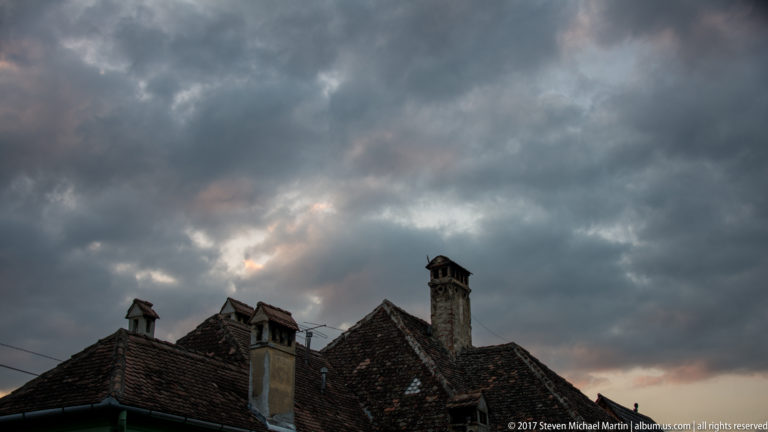 Weathered red-brown tile roofs and tall masonry chimneys silhouetted beneath dark, dramatic storm clouds in Sighisoara.