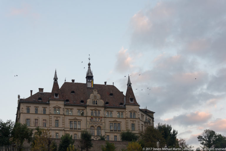 Large symmetrical Sighisoara City Hall with steep tiled roof, central entrance bay, dormers, and slender corner turrets beneath cloudy sky.