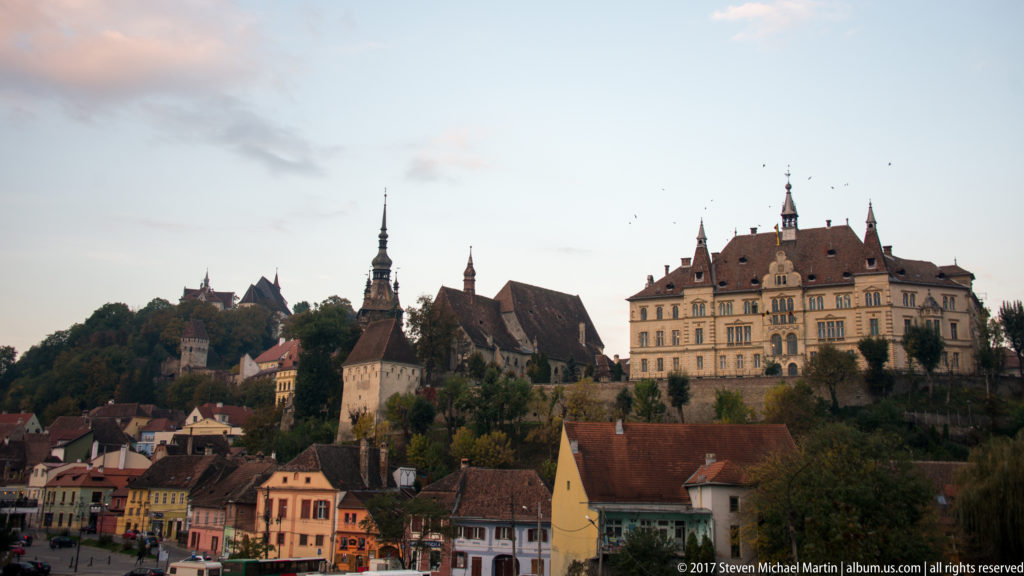 Sighisoara citadel hill rising above red-tiled houses, with towers, church roofs, and a large historic civic building on the ridge.