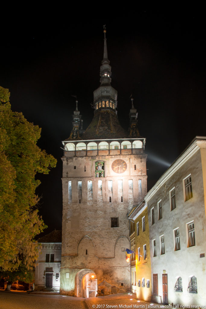 Illuminated Sighisoara Clock Tower with arched gateway and adjoining historic buildings at night.