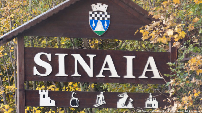 Wooden SINAIA sign with a heraldic crest and white landmark pictograms, surrounded by autumn foliage.