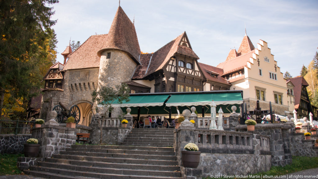 Stone steps rise toward a terrace with canopy seating; a round tower, arched gateway, half-timbered gable, and steep roofs form the backdrop