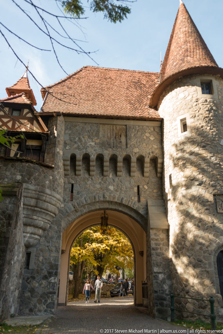 Broad masonry arch passes through a crenellated-looking wall beside a round stone tower capped by a steep conical tile roof