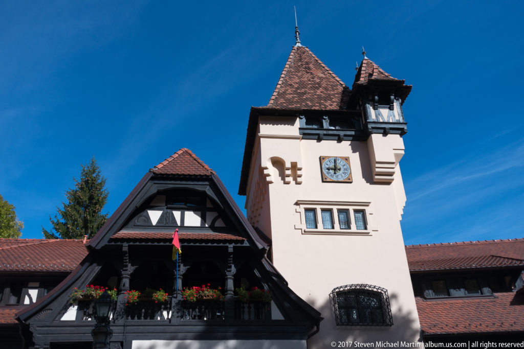 Pale square clock tower with red tile roof rises beside a dark timber gable, balcony flower boxes, and Romanian flag