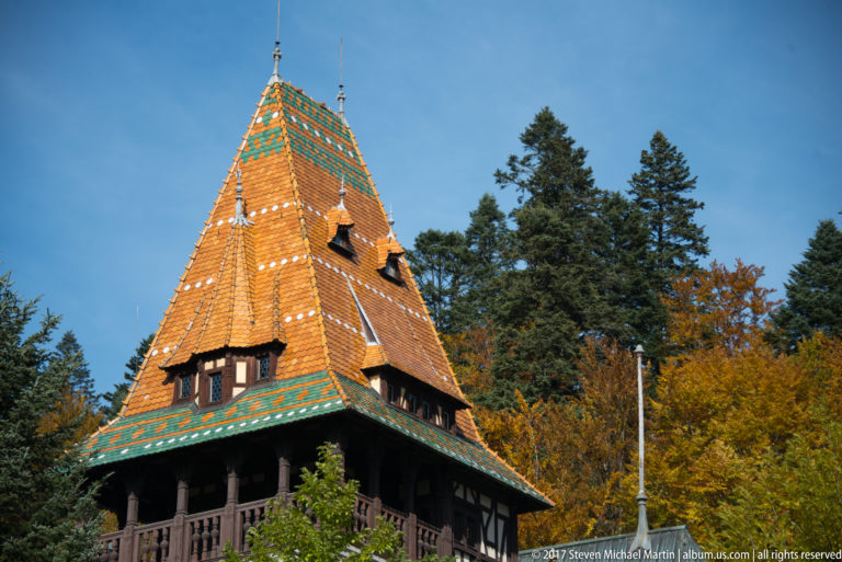 Steep orange tile roof carries green geometric bands and small dormers above a dark timber gallery, backed by conifers and autumn trees