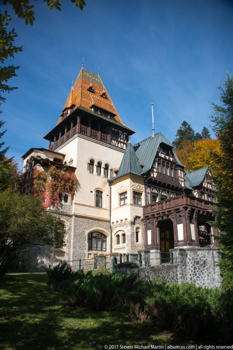 Wide side view shows the orange-roofed tower, pale masonry base, dark timber wings, covered porch, lawn, and wooded hillside
