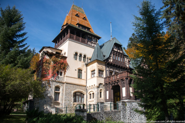 Three-quarter view frames a tall square tower with patterned roof, pale stucco and stone base, adjoining dark timber galleries, and evergreen trees