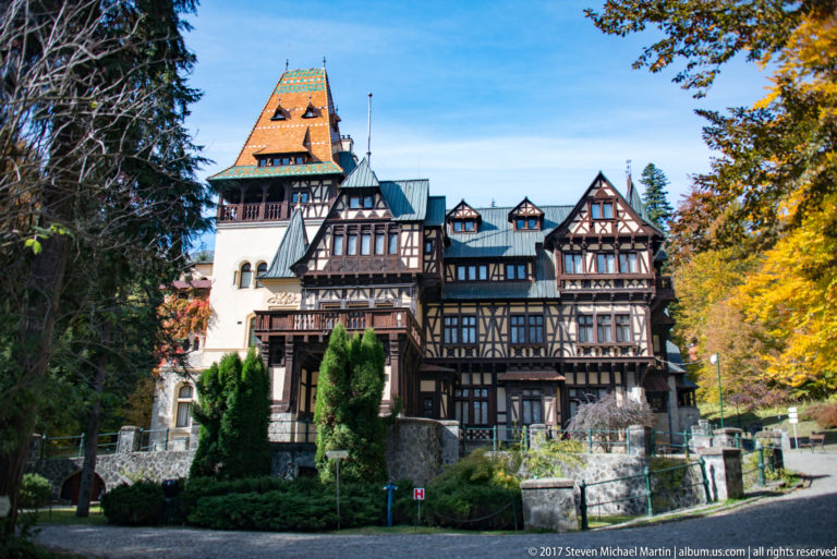 Broad frontal view shows a multiwing villa with dark half-timbering, pale masonry, projecting balconies, and a tall patterned orange-and-green roof