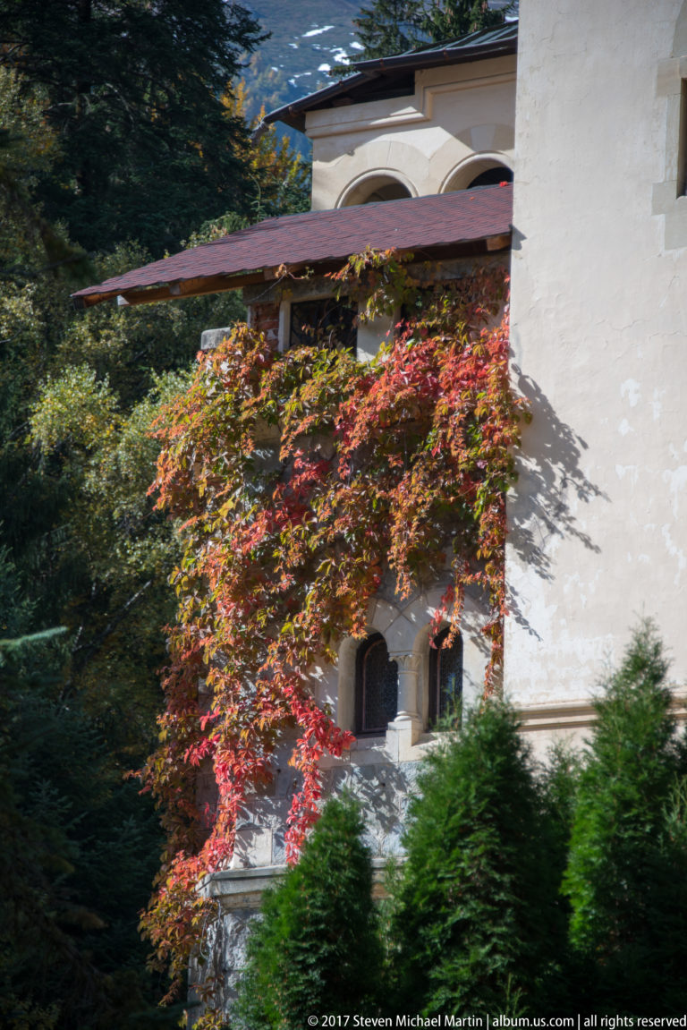 White plaster wall with small arched openings and a broad red-tiled eave is partly covered by dense red autumn vine
