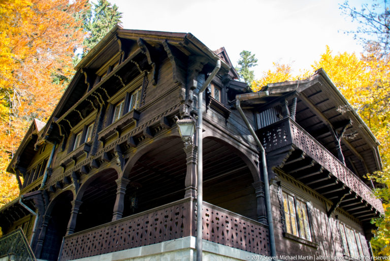 Low-angle view shows a dark wooden building with deep eaves, carved brackets, wraparound balconies, arched supports, and autumn trees