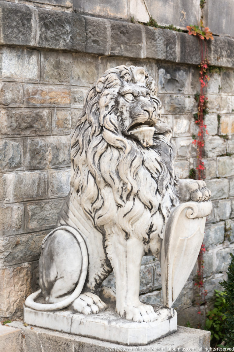 Carved seated lion with full mane rests one paw on a shield against a rough stone wall with sparse red vine