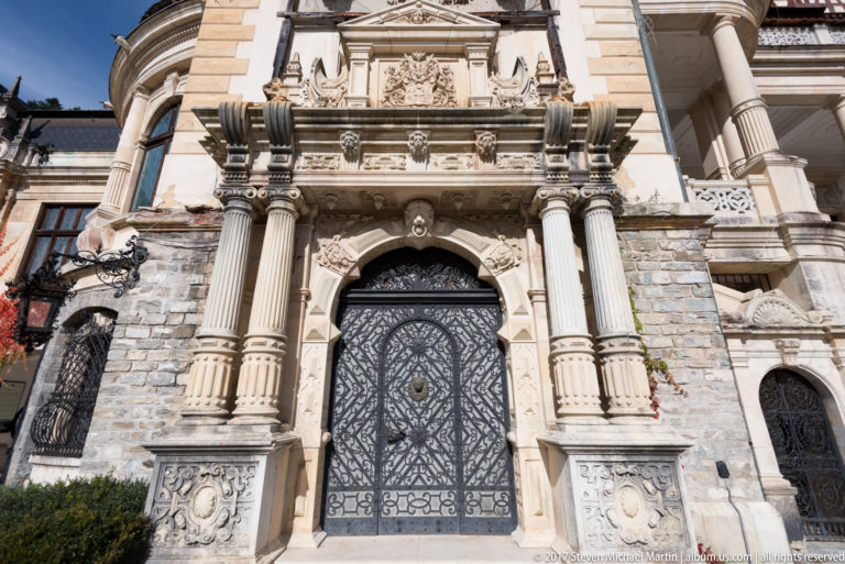 Tall patterned wrought-iron door sits within a richly carved stone portal with paired columns, sculpted capitals, relief panels, and heraldic ornament