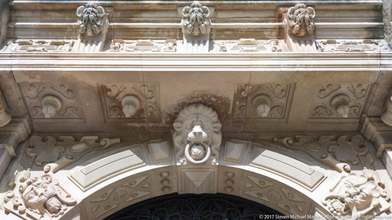 Upward close view of an arched entrance shows a central carved mask holding a ring, coffering, floral reliefs, and projecting cornice