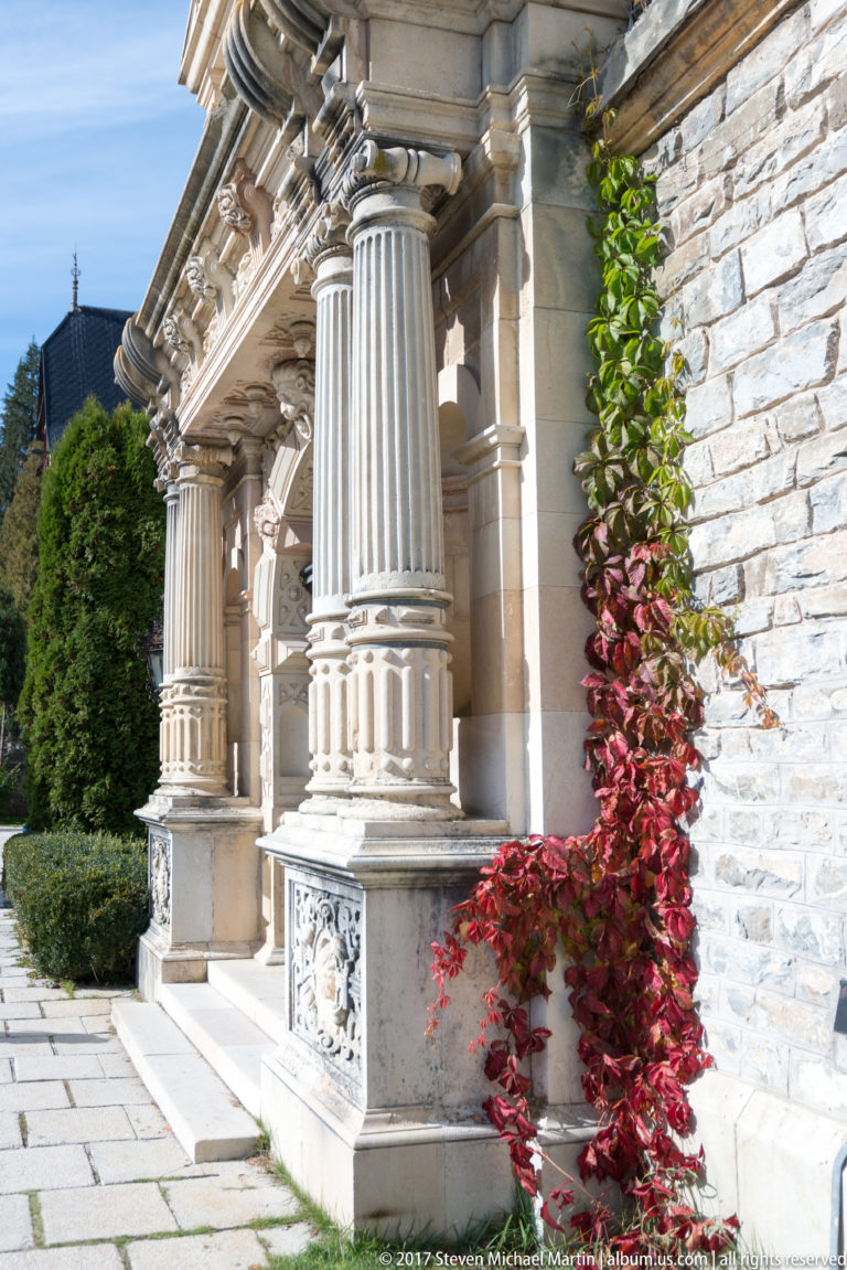 Close oblique view shows paired fluted columns, carved bases and entablature against rough stone masonry, with red climbing leaves