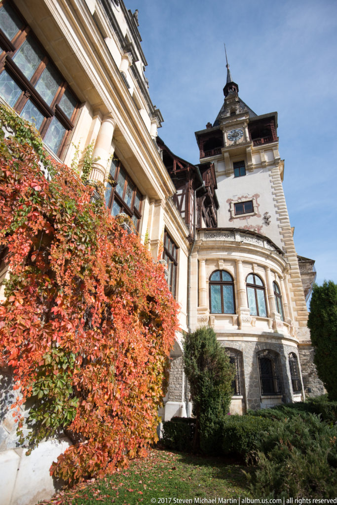 Cream masonry wing with arched windows, carved ornament, dark timber roof structures, and tower rises behind brilliant red foliage