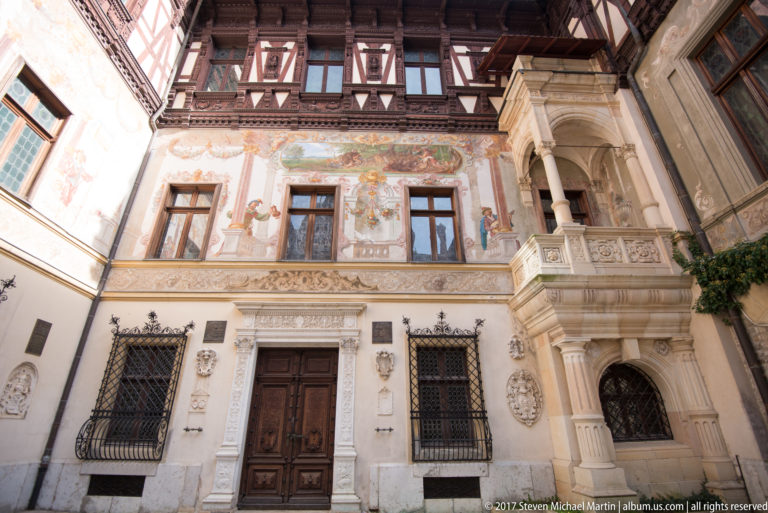 Frontal courtyard wall combines mural-like painted ornament, carved stone balcony, timber-framed upper story, grilled windows, and central wooden door