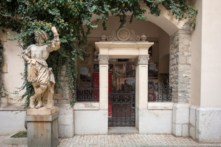 Standing carved figure flanks an arched opening containing a small classical stone doorway, iron gate, red carpet, and ivy