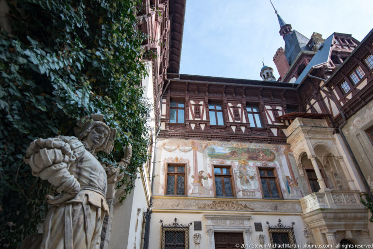 Stone figure in foreground stands against ivy; behind it are painted plaster walls, timber framing, windows, and a carved balcony