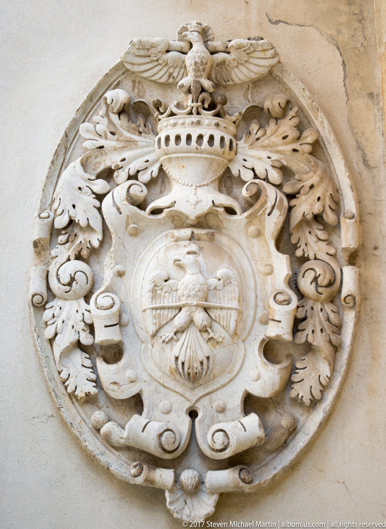 Oval stone cartouche contains a shield and eagle-like heraldic device beneath a crown, surrounded by scrolling foliage
