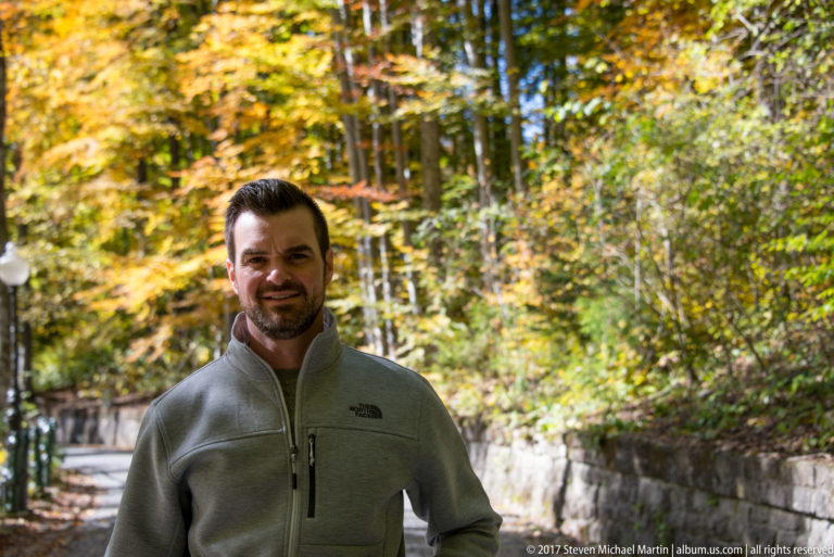 Waist-up portrait of a man in a gray-green zip jacket standing on a paved path bordered by stone wall and yellow autumn woodland