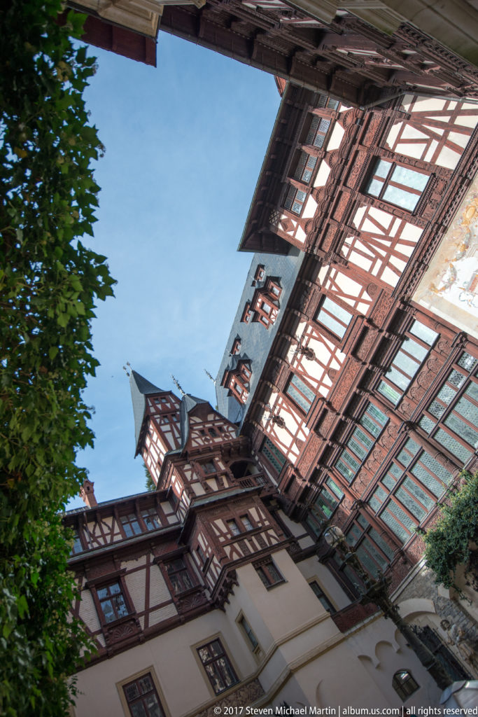 Steep upward view encloses half-timbered upper stories, projecting bays, painted wall decoration, windows, and blue sky